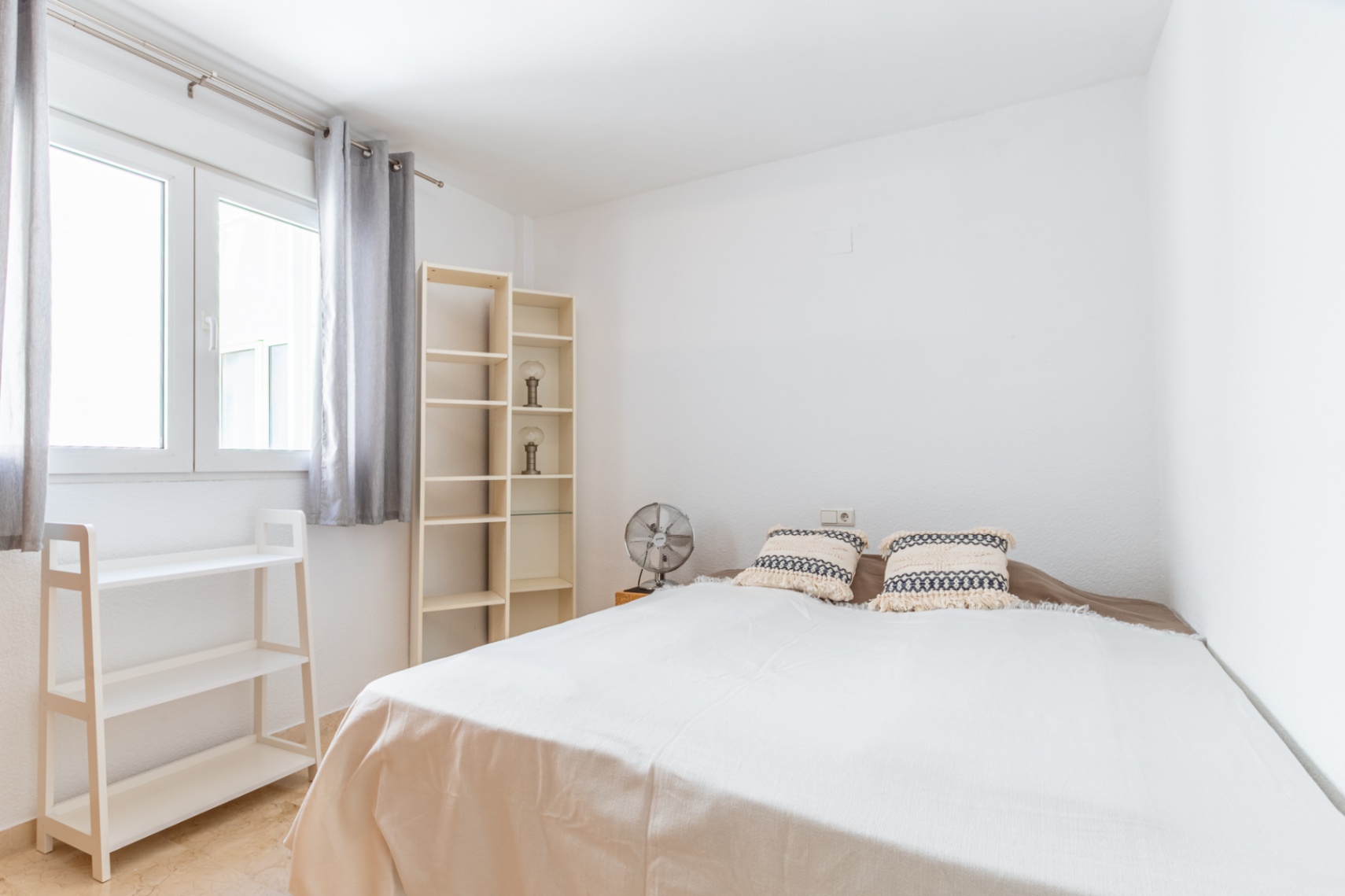 Dormitorio luminoso con cama doble, estanterías de madera clara y ventilador. La ventana con cortinas grises aporta abundante luz natural al espacio de paredes blancas y suelo de mármol. Dormitorio luminoso con cama doble, estanterías de madera clara y ventilador. La ventana con cortinas grises aporta abundante luz natural al espacio de paredes blancas y suelo de mármol.
