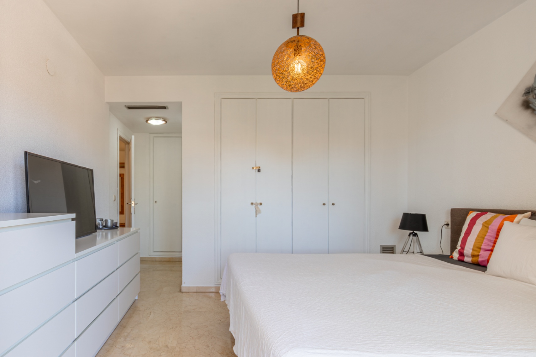 Dormitorio principal con armarios empotrados blancos, suelo de mármol claro y una lámpara colgante esférica. Incluye cómoda con TV y acceso a pasillo. Dormitorio principal con armarios empotrados blancos, suelo de mármol claro y una lámpara colgante esférica. Incluye cómoda con TV y acceso a pasillo.