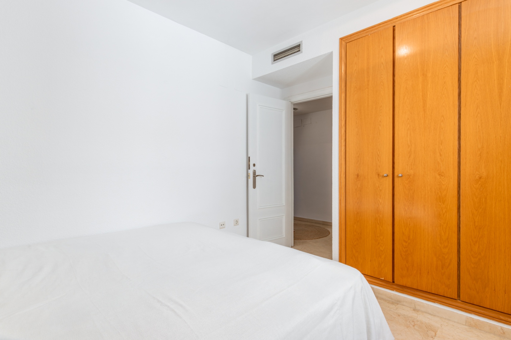 Dormitorio secundario con amplio armario empotrado de madera clara y cama con ropa blanca. Destaca el suelo de mármol y la puerta abierta que conecta a otra estancia. Dormitorio secundario con amplio armario empotrado de madera clara y cama con ropa blanca. Destaca el suelo de mármol y la puerta abierta que conecta a otra estancia.