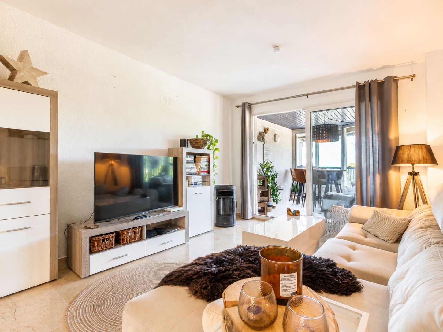 Luminoso salón con sofá seccional claro, mesa de centro blanca brillante y mueble de TV. Puertas correderas de cristal conectan con una terraza cubierta, ampliando el área social. Luminoso salón con sofá seccional claro, mesa de centro blanca brillante y mueble de TV. Puertas correderas de cristal conectan con una terraza cubierta, ampliando el área social.