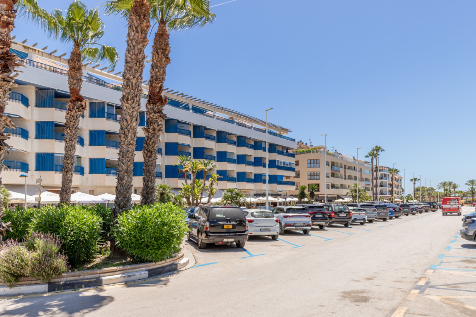Vista exterior de una calle costera con un edificio de varios pisos y balcones azules, palmeras y coches aparcados bajo un cielo despejado. Vista exterior de una calle costera con un edificio de varios pisos y balcones azules, palmeras y coches aparcados bajo un cielo despejado.