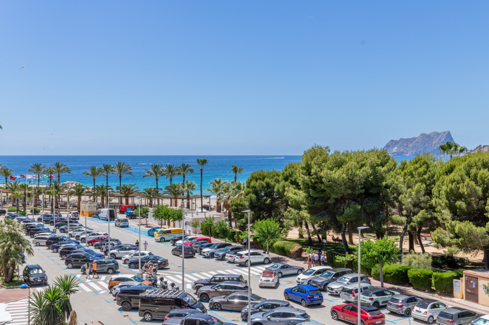 Vista panorámica al mar Mediterráneo, con palmeras, playa de arena y el Peñón de Ifach a lo lejos, junto a una animada zona de aparcamiento y pinos. Vista panorámica al mar Mediterráneo, con palmeras, playa de arena y el Peñón de Ifach a lo lejos, junto a una animada zona de aparcamiento y pinos.