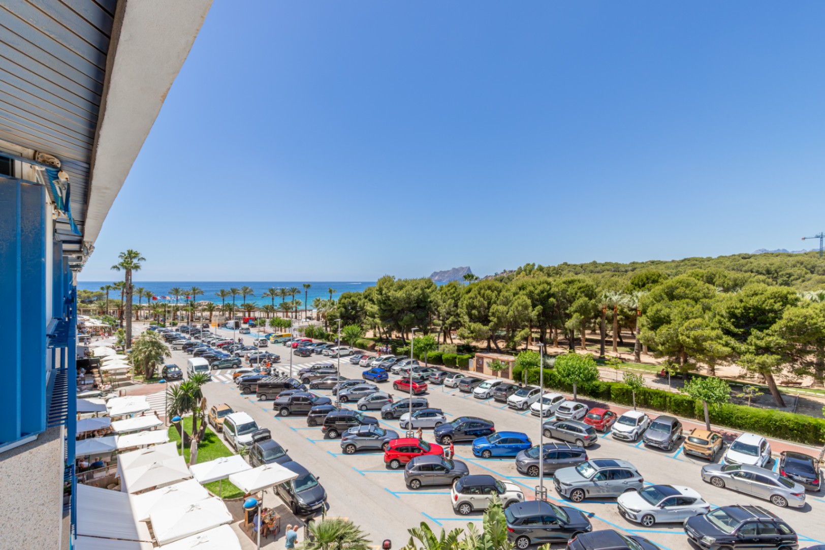 Vista panorámica desde la terraza, con el mar Mediterráneo, una playa de palmeras y el Peñón de Ifach en el horizonte. Abajo, un aparcamiento y terrazas con toldos blancos. Vista panorámica desde la terraza, con el mar Mediterráneo, una playa de palmeras y el Peñón de Ifach en el horizonte. Abajo, un aparcamiento y terrazas con toldos blancos.