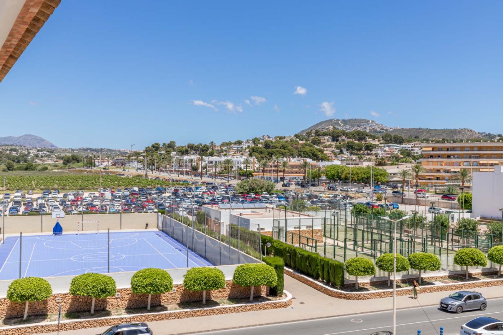 Vista panorámica que incluye una cancha deportiva azul, varias pistas de pádel, un amplio aparcamiento y el paisaje urbano de Moraira con montañas al fondo. Vista panorámica que incluye una cancha deportiva azul, varias pistas de pádel, un amplio aparcamiento y el paisaje urbano de Moraira con montañas al fondo.