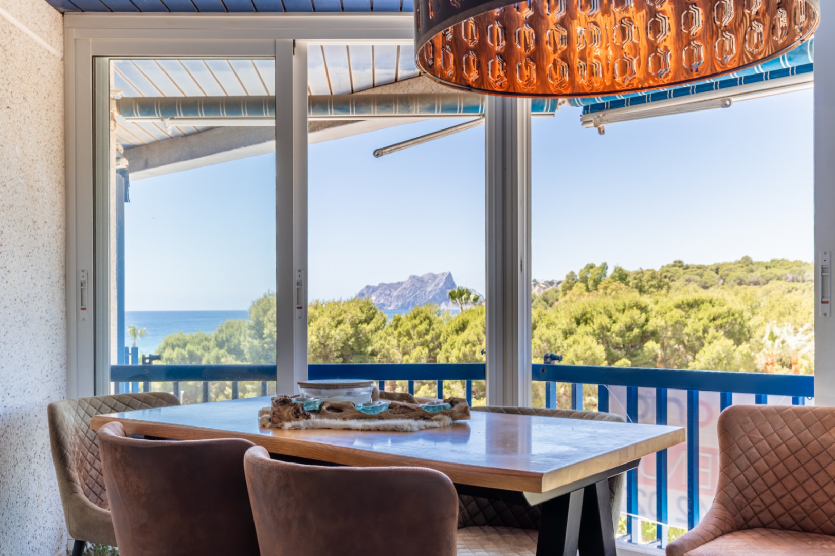 Zona de comedor con vistas panorámicas al mar, montaña y exuberante vegetación a través de amplias puertas correderas de cristal. Destaca una lámpara colgante decorativa única y sillas tapizadas. Zona de comedor con vistas panorámicas al mar, montaña y exuberante vegetación a través de amplias puertas correderas de cristal. Destaca una lámpara colgante decorativa única y sillas tapizadas.