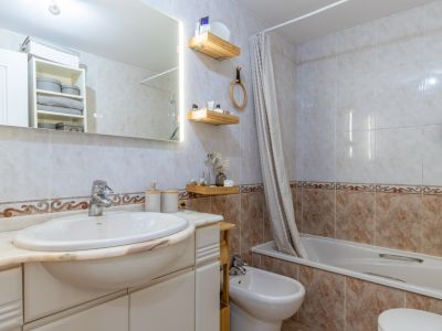 Baño completo con azulejos claros y cenefa decorativa, lavabo integrado en encimera de mármol, espejo retroiluminado, bañera con cortina y bidé.