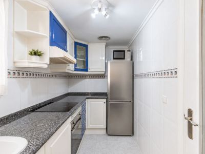 Cocina funcional con armarios blancos y azules, encimeras de granito oscuro, placa de inducción integrada y electrodomésticos de acero inoxidable. Iluminación natural junto a la ventana.