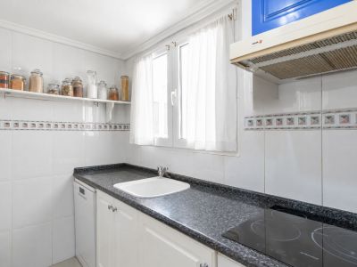 Cocina independiente con encimera de granito oscuro, fregadero blanco y placa de inducción. Paredes alicatadas en blanco con cenefa decorativa y ventana con luz natural.