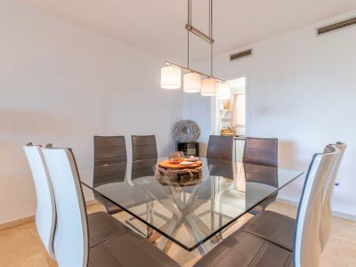 Comedor con mesa cuadrada de cristal y base metálica, rodeada de sillas de cuero. Una lámpara colgante moderna ilumina el espacio, que conecta con la cocina.