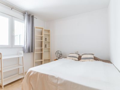 Dormitorio luminoso con cama doble, estanterías de madera clara y ventilador. La ventana con cortinas grises aporta abundante luz natural al espacio de paredes blancas y suelo de mármol.