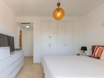 Dormitorio principal con armarios empotrados blancos, suelo de mármol claro y una lámpara colgante esférica. Incluye cómoda con TV y acceso a pasillo.