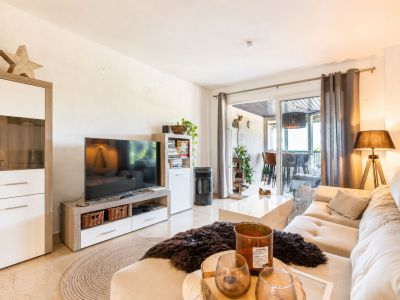 Luminoso salón con sofá seccional claro, mesa de centro blanca brillante y mueble de TV. Puertas correderas de cristal conectan con una terraza cubierta, ampliando el área social.