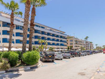Vista exterior de una calle costera con un edificio de varios pisos y balcones azules, palmeras y coches aparcados bajo un cielo despejado.