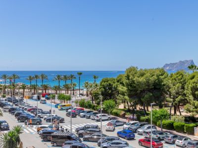 Vista panorámica al mar Mediterráneo, con palmeras, playa de arena y el Peñón de Ifach a lo lejos, junto a una animada zona de aparcamiento y pinos.