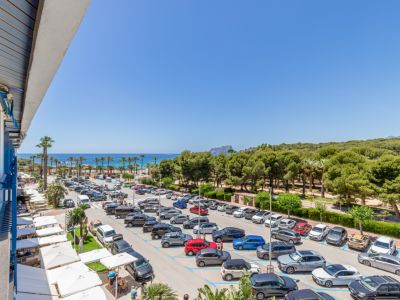 Vista panorámica desde la terraza, con el mar Mediterráneo, una playa de palmeras y el Peñón de Ifach en el horizonte. Abajo, un aparcamiento y terrazas con toldos blancos.