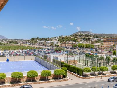 Vista panorámica que incluye una cancha deportiva azul, varias pistas de pádel, un amplio aparcamiento y el paisaje urbano de Moraira con montañas al fondo.