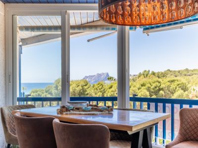 Zona de comedor con vistas panorámicas al mar, montaña y exuberante vegetación a través de amplias puertas correderas de cristal. Destaca una lámpara colgante decorativa única y sillas tapizadas.
