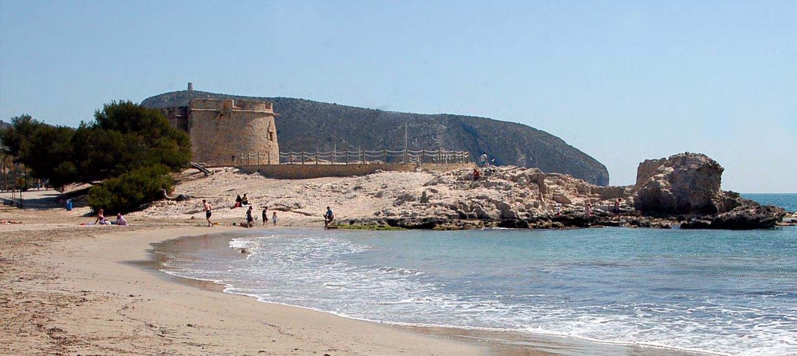 Playa de arena con una torre de vigilancia de piedra y formaciones rocosas costeras, bañada por el mar Mediterráneo bajo un cielo azul despejado.