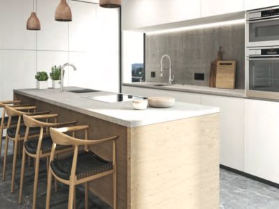 Cocina moderna con isla de hormigón claro, placa de inducción y fregadero integrados, taburetes de madera y armarios blancos sin tiradores. Iluminación empotrada en el salpicadero de hormigón.