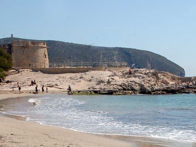 Playa de arena con una torre de vigilancia de piedra y formaciones rocosas costeras, bañada por el mar Mediterráneo bajo un cielo azul despejado.
