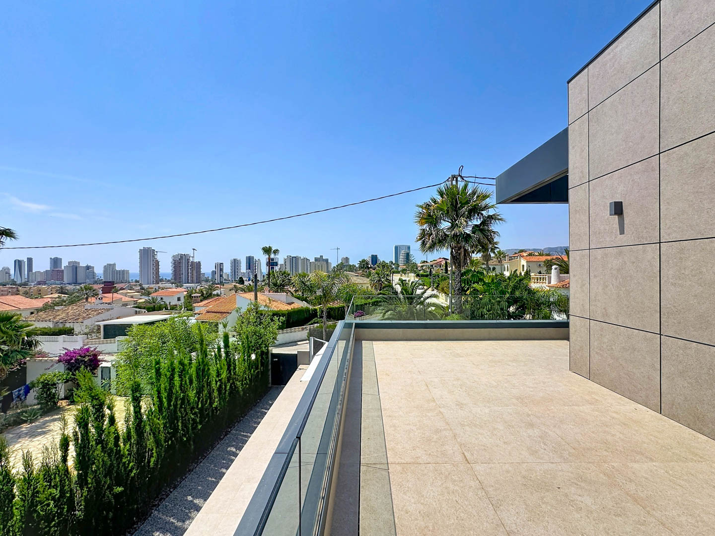 Amplia terraza superior con suelo de gres claro y barandilla de cristal, ofreciendo vistas panorámicas al skyline de Calpe, el mar y la vegetación mediterránea. La fachada moderna de paneles completa el diseño.