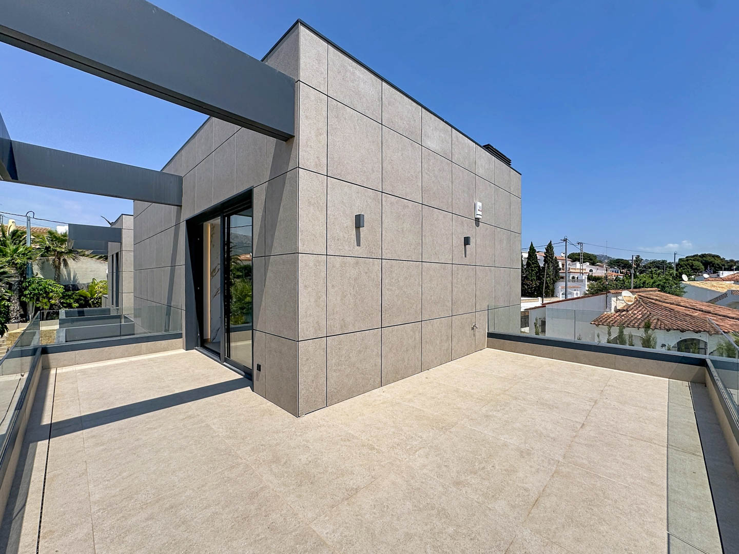 Amplia terraza superior con suelo porcelánico y barandillas de cristal, bajo vigas estructurales. La fachada panelada moderna se abre a vistas residenciales.