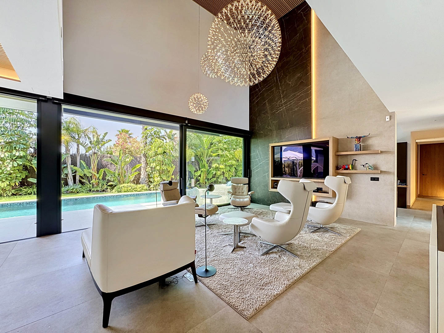 Amplio salón de doble altura con pared de acento de efecto mármol oscuro, elegantes lámparas esféricas y grandes puertas de cristal que se abren a una piscina privada y jardín tropical.