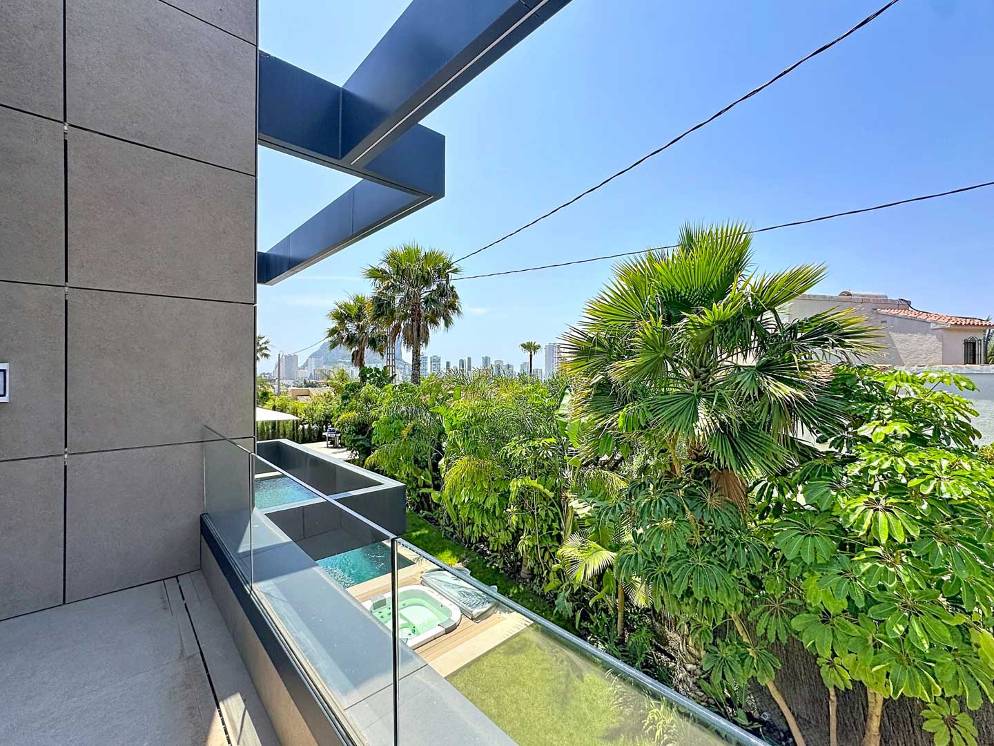 Balcón moderno con fachada de paneles grises y barandilla de cristal, revelando jacuzzis exteriores, exuberante vegetación tropical y el skyline de Calpe al fondo.