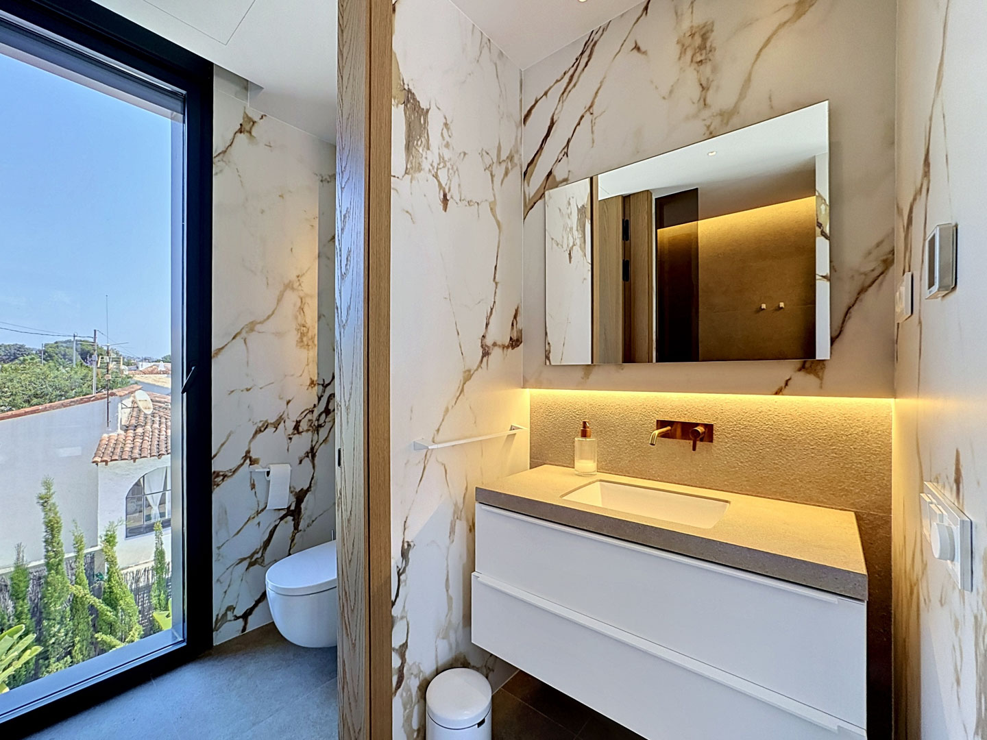 Baño de diseño con revestimiento mural de mármol veteado y un gran ventanal que ofrece vistas residenciales. Incluye tocador flotante con encimera texturizada y grifería empotrada.