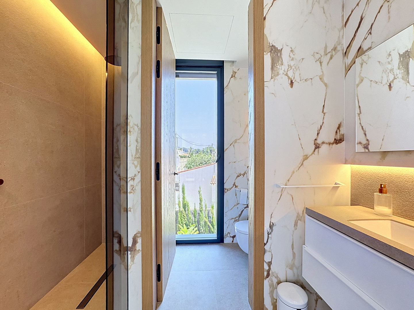 Baño moderno con ducha a ras de suelo en tonos beige y elegantes paredes de mármol veteado. El tocador flotante incorpora iluminación, y una ventana de altura completa ofrece vistas al exterior.