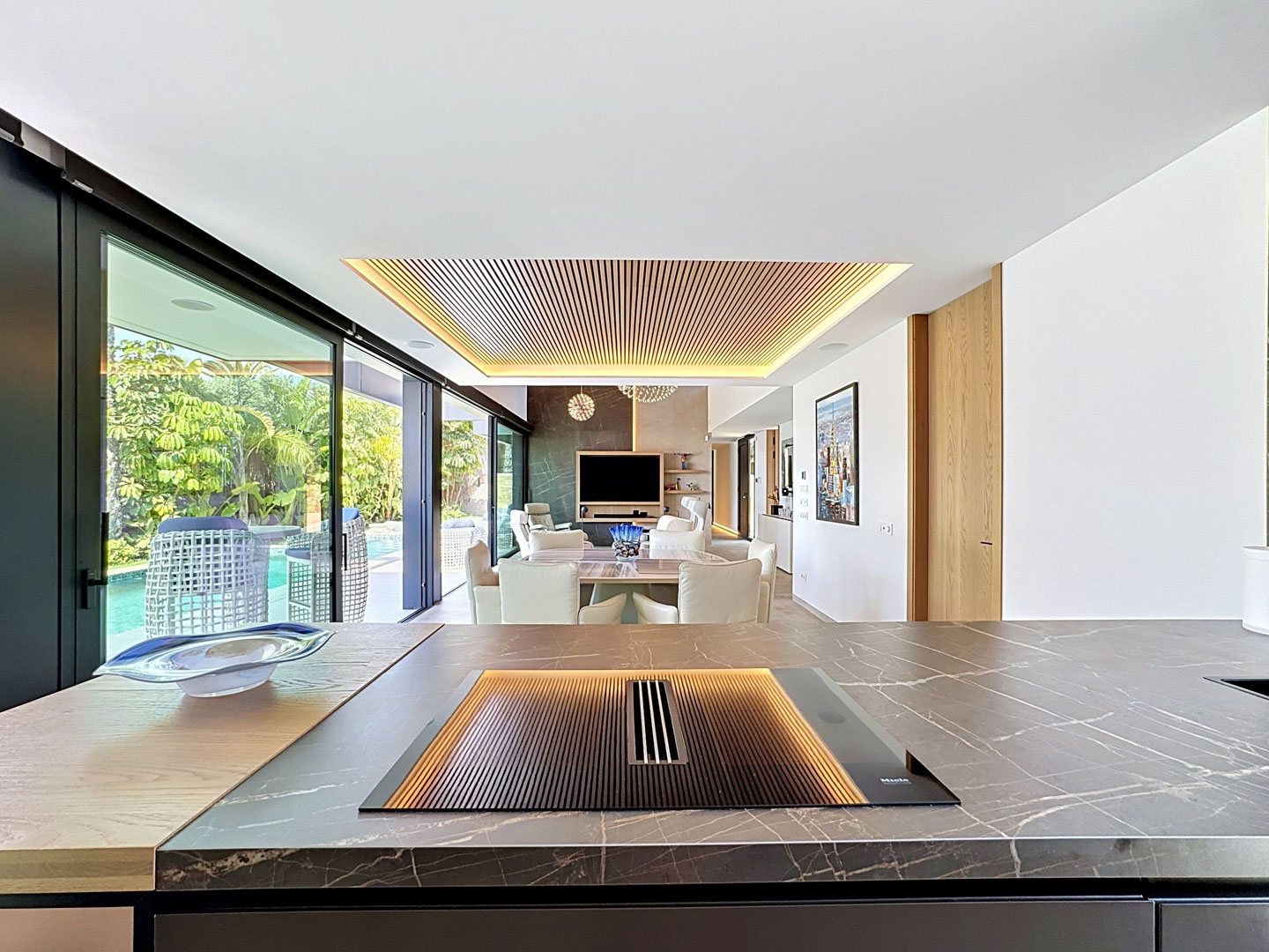 Cocina de diseño con encimera de mármol veteado y placa Miele integrada, abierta al salón con techo de lamas de madera iluminado y acceso directo a la piscina y jardines.