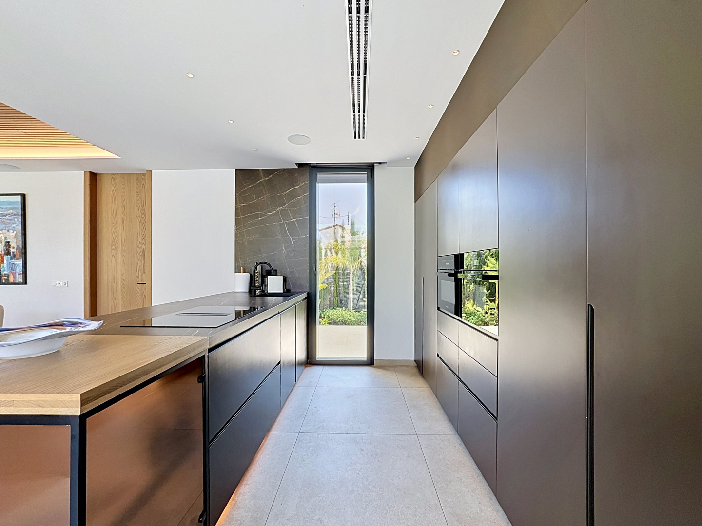 Cocina de diseño con mobiliario oscuro sin tiradores y electrodomésticos Miele integrados. La isla central cuenta con encimera negra, placa y fregadero, complementada por una pared de piedra veteada y salida al jardín.