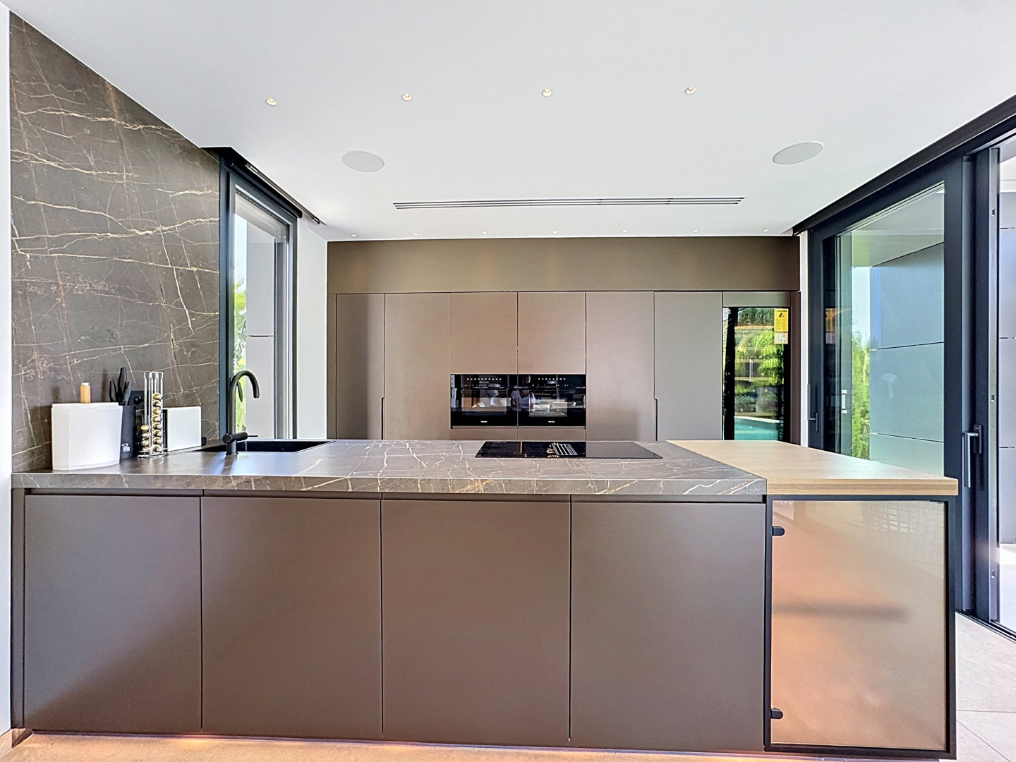 Cocina moderna con isla central de encimera de piedra veteada, fregadero integrado y placa de inducción. Hornos Miele empotrados en la ebanistería minimalista, con vinoteca y acceso directo al exterior.