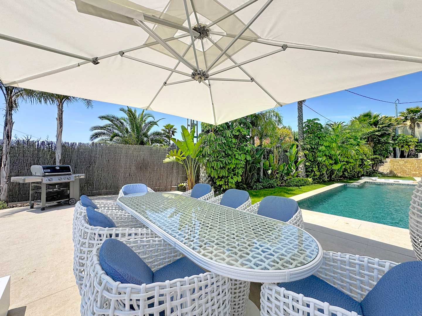 Comedor exterior bajo una gran sombrilla blanca, con mesa de cristal y sillas trenzadas blancas con cojines azules, junto a la piscina y un jardín tropical.