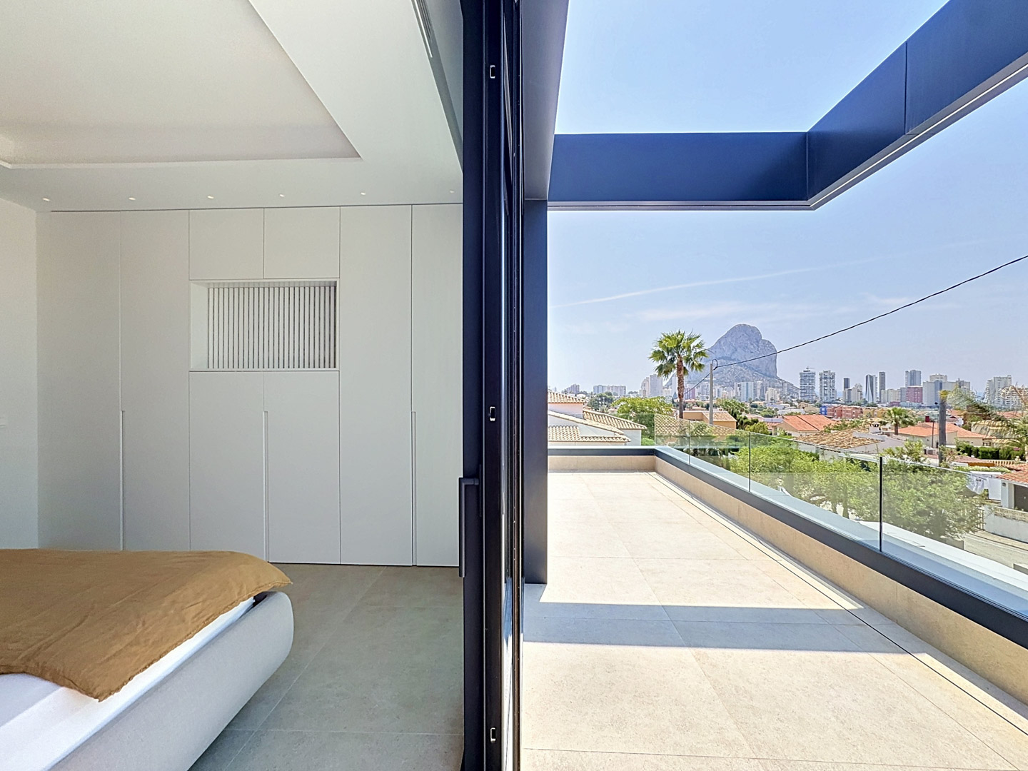 Dormitorio con armarios empotrados blancos y cama moderna, con acceso directo a una terraza exterior. Disfruta de vistas panorámicas a Calpe y al majestuoso Peñón de Ifach.