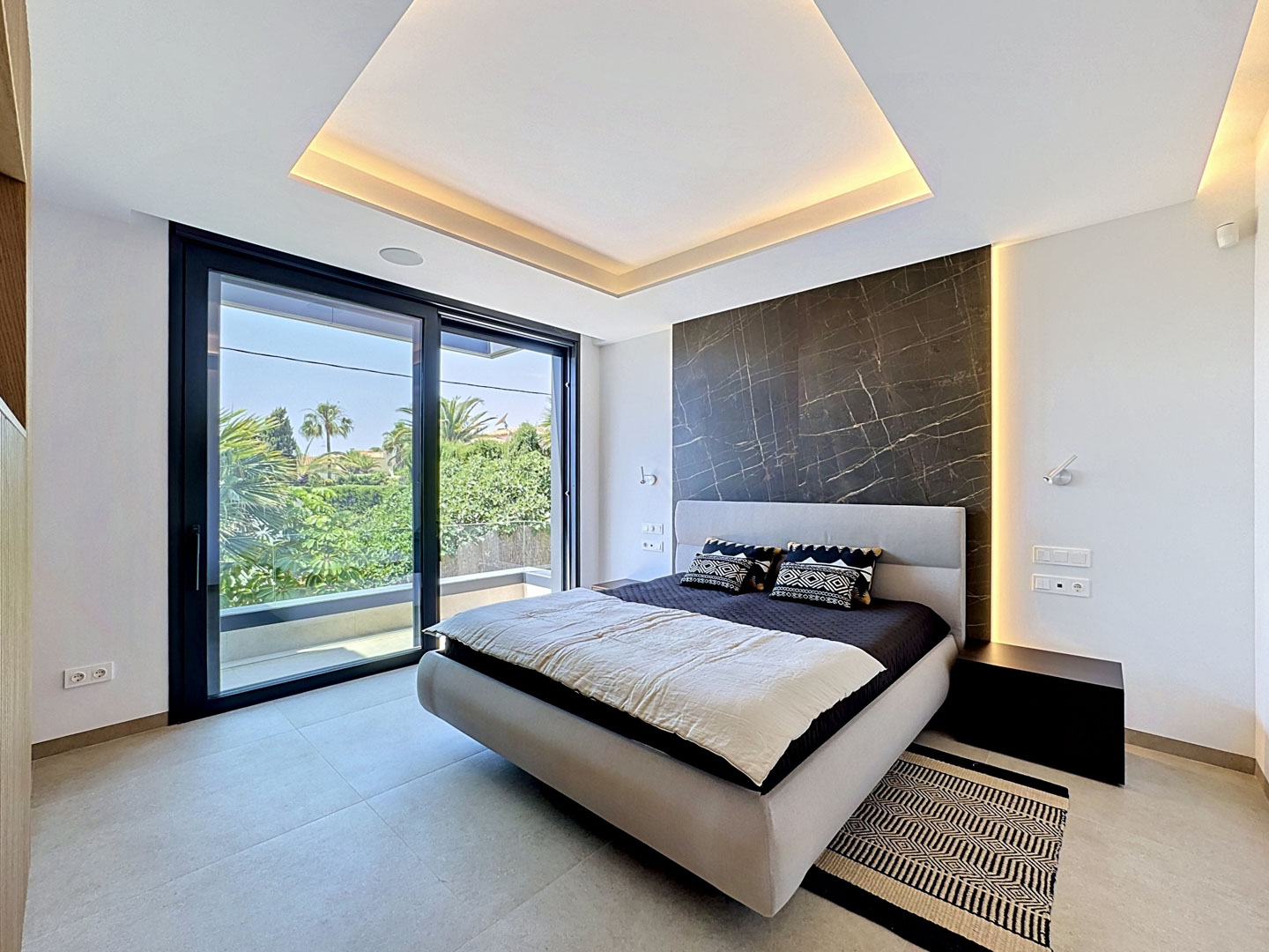 Dormitorio con cama de diseño, panel de mármol oscuro y cabecero tapizado. El ventanal corredero ofrece vistas a la vegetación exterior y acceso a la terraza, complementado por iluminación LED integrada en techo y pared.