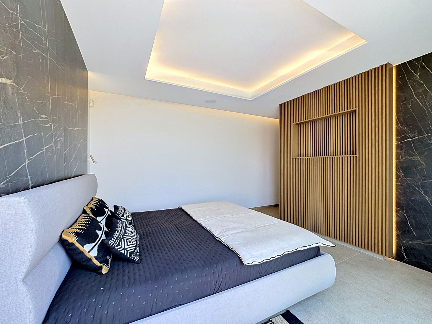 Dormitorio moderno con cama tapizada en gris claro, paneles de pared efecto mármol oscuro y una pared central de lamas de madera con iluminación integrada y estante empotrado. El techo presenta iluminación perimetral.