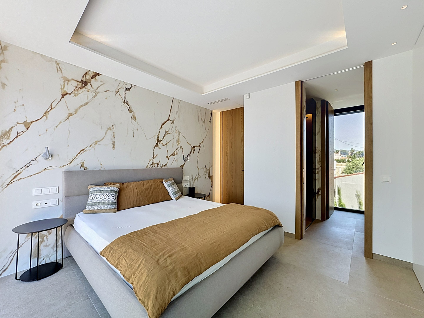 Dormitorio principal con cabecero de tela, pared revestida en porcelánico efecto mármol con vetas doradas y detalles de iluminación indirecta en techo. Acceso a zona de vestidor/baño y exterior.