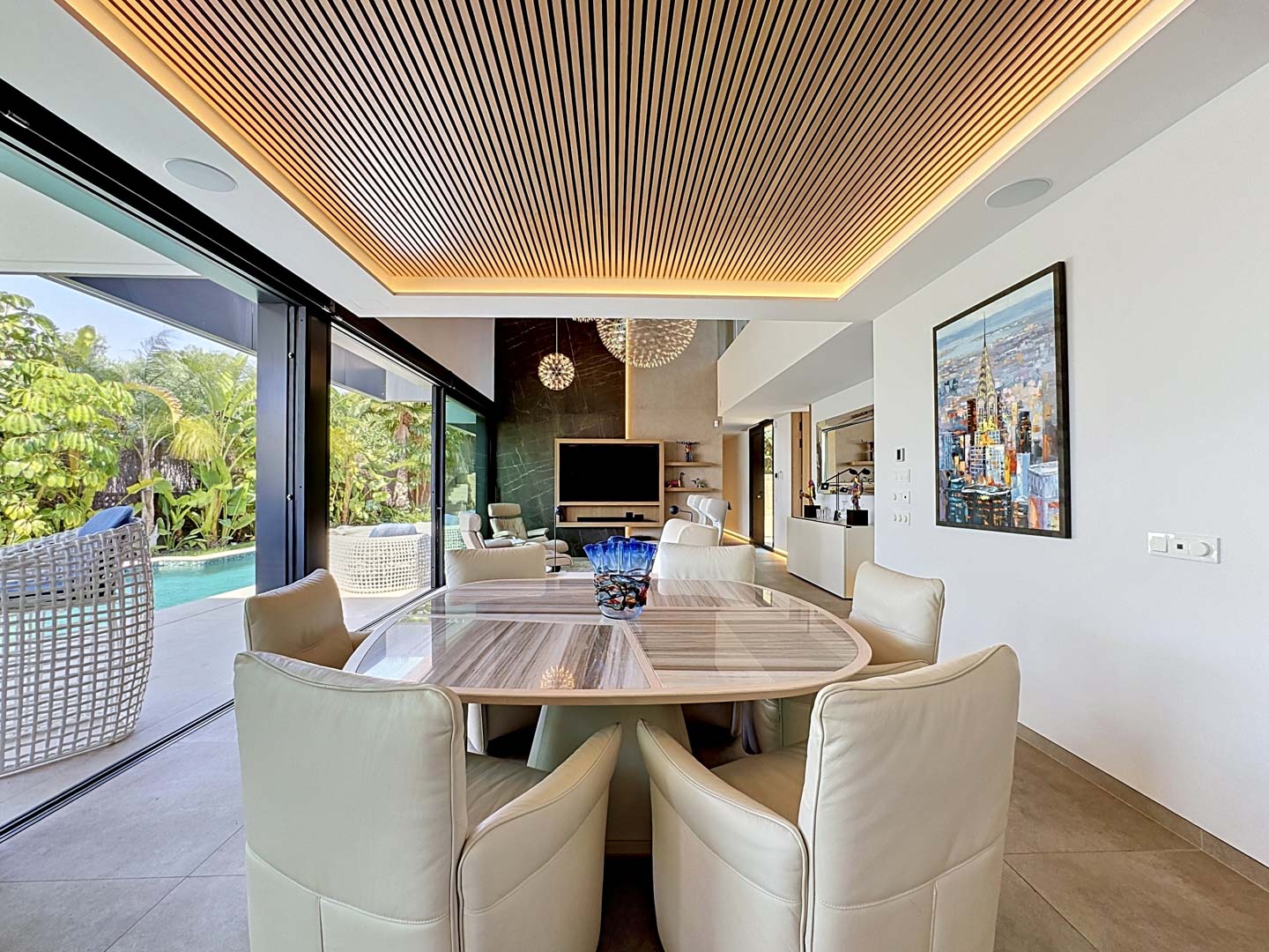 Elegante comedor con mesa ovalada y sillas tapizadas, destacando un techo de lamas de madera iluminado y acceso directo a la piscina y jardines tropicales.