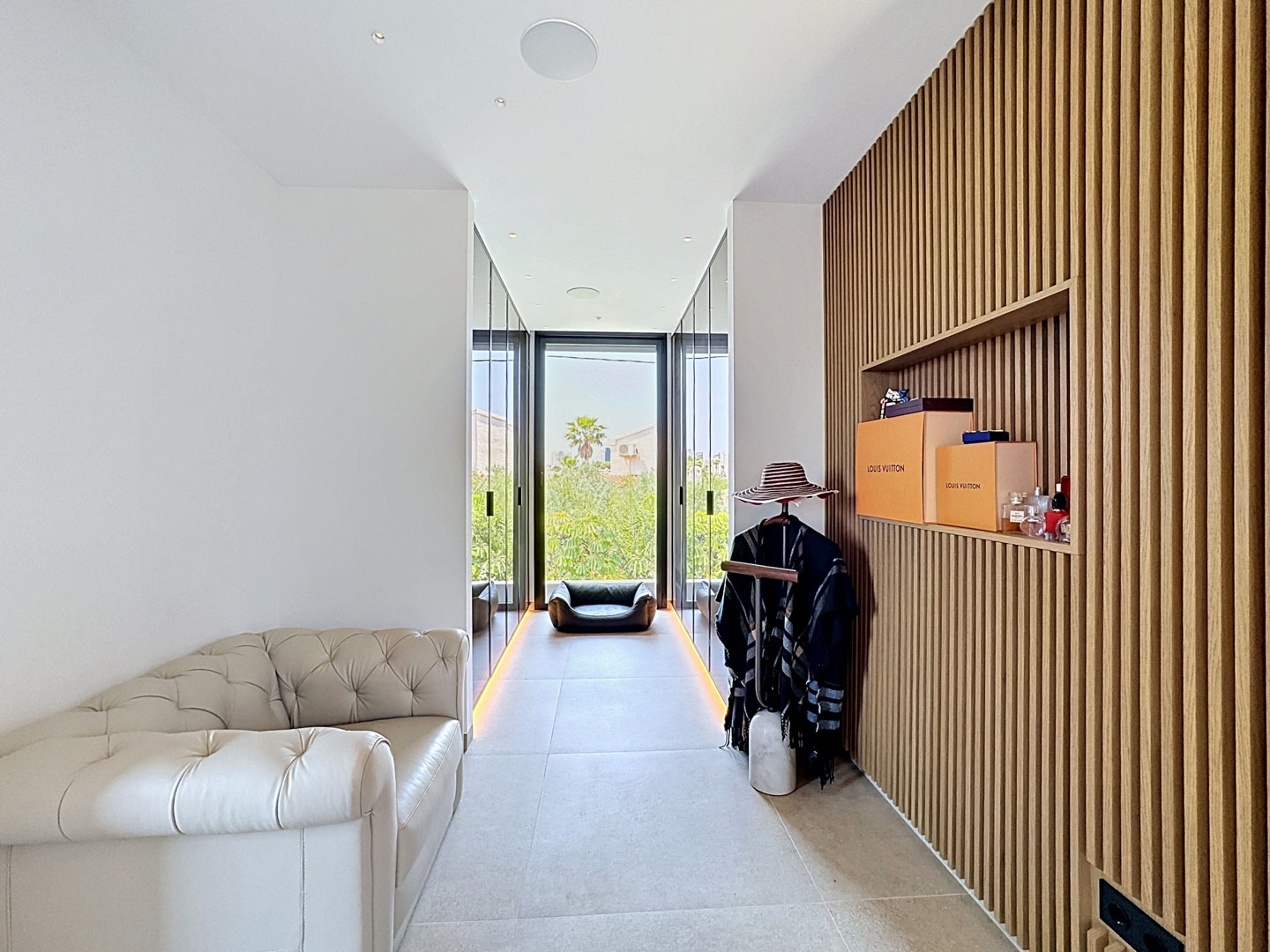 Elegante zona de vestidor con pared de lamas de madera y estantería integrada, un sofá clásico de cuero capitoné y grandes puertas de cristal hacia jardines.