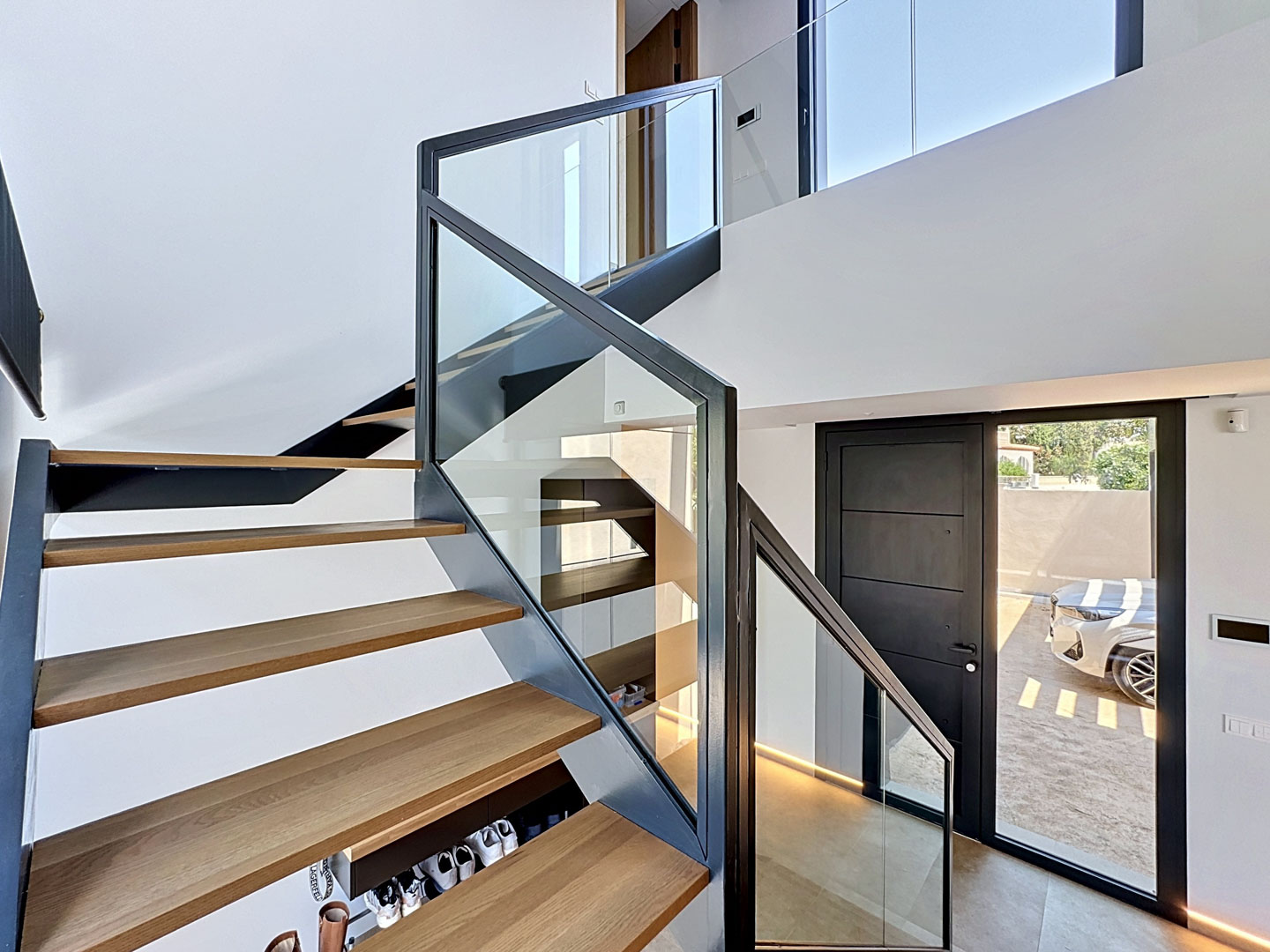 Escalera flotante contemporánea con peldaños de madera de roble y barandilla de cristal, iluminada por luz natural en un vestíbulo de doble altura.
