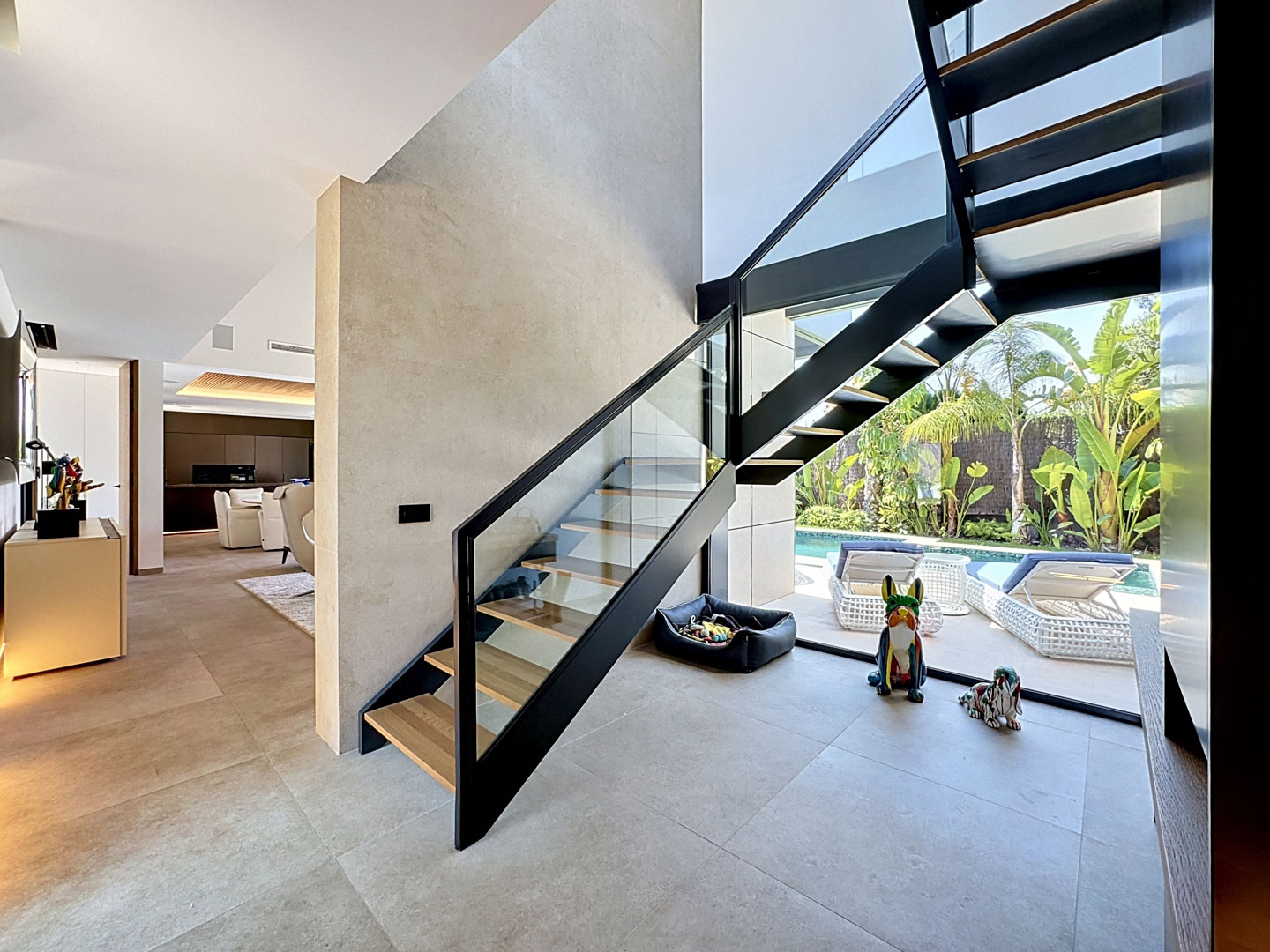 Escalera moderna de peldaños abiertos con barandilla de cristal y metal negro, conectando interiores diáfanos y vistas a la piscina privada con jardín tropical.