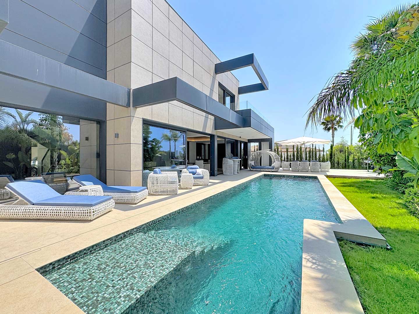 Exterior de villa contemporánea con piscina privada de mosaico, rodeada por una terraza de baldosas claras y mobiliario lounge de ratán blanco. Jardines tropicales exuberantes y césped cuidado completan este oasis.