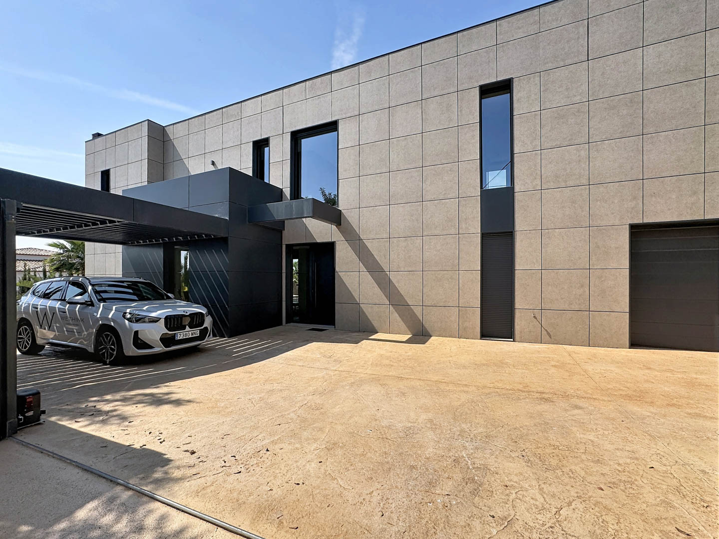 Fachada minimalista de la villa con revestimiento de paneles claros y entrada en antracita. El porche de lamas metálicas protege un vehículo, junto a un amplio acceso pavimentado y garaje.