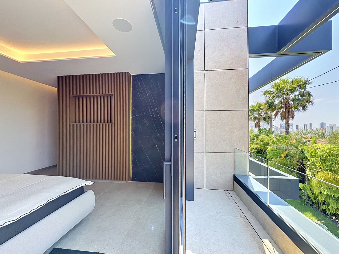 Interior de dormitorio con cabecero de lamas de madera y panel de mármol retroiluminado. Acceso a balcón con barandilla de cristal y vistas a palmeras y la ciudad.