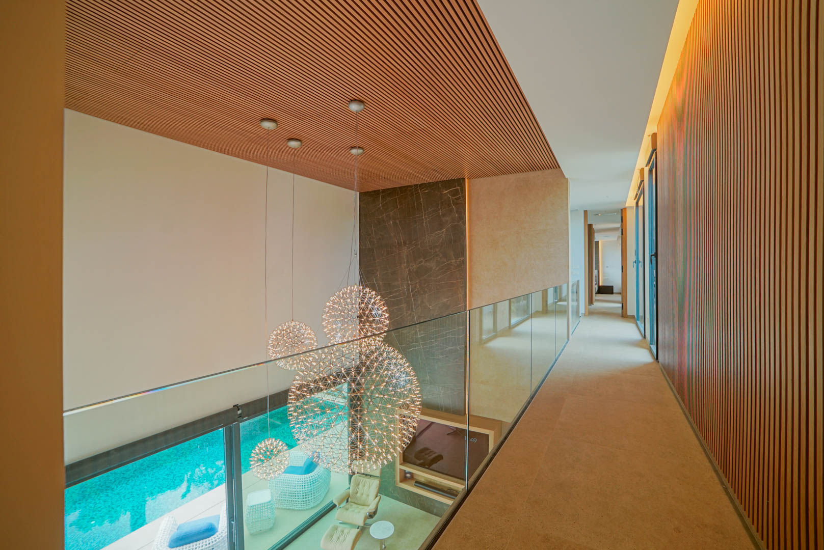 Pasillo superior con barandilla de cristal y techo de lamas de madera, ofreciendo vistas a un salón de doble altura con lámparas esféricas y acceso a la piscina.