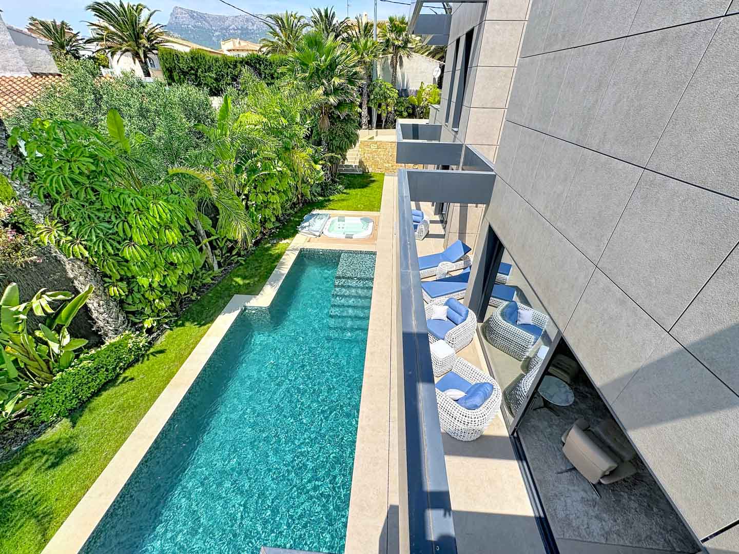 Piscina rectangular con jacuzzi integrado, rodeada de exuberantes jardines tropicales y césped. Balcones modernos con mobiliario de exterior y fachada de baldosas grises.