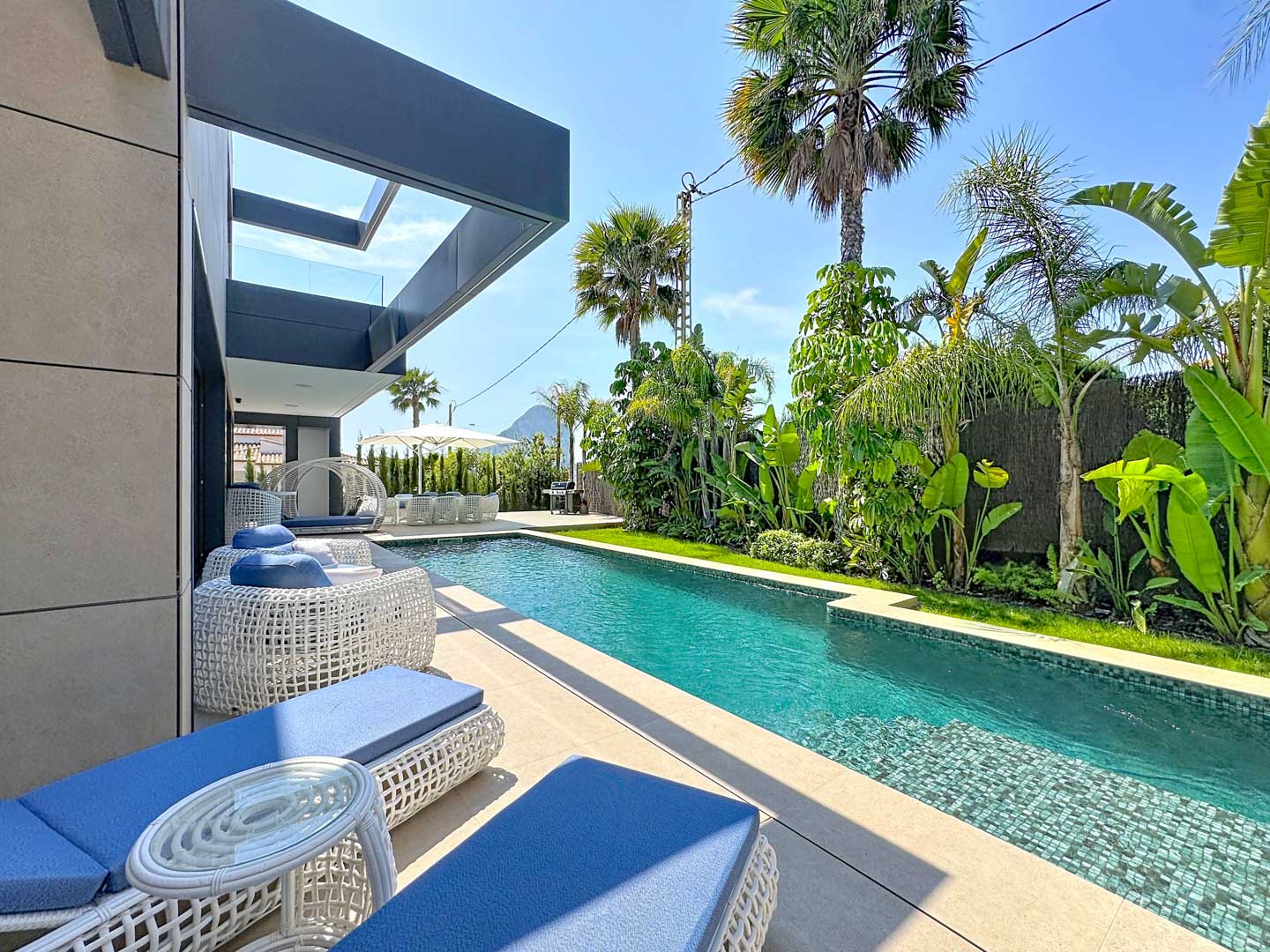 Piscina rectangular con mosaico azul, rodeada de tumbonas blancas con cojines azules y exuberante vegetación tropical. La arquitectura moderna de la villa se integra con el espacio exterior.