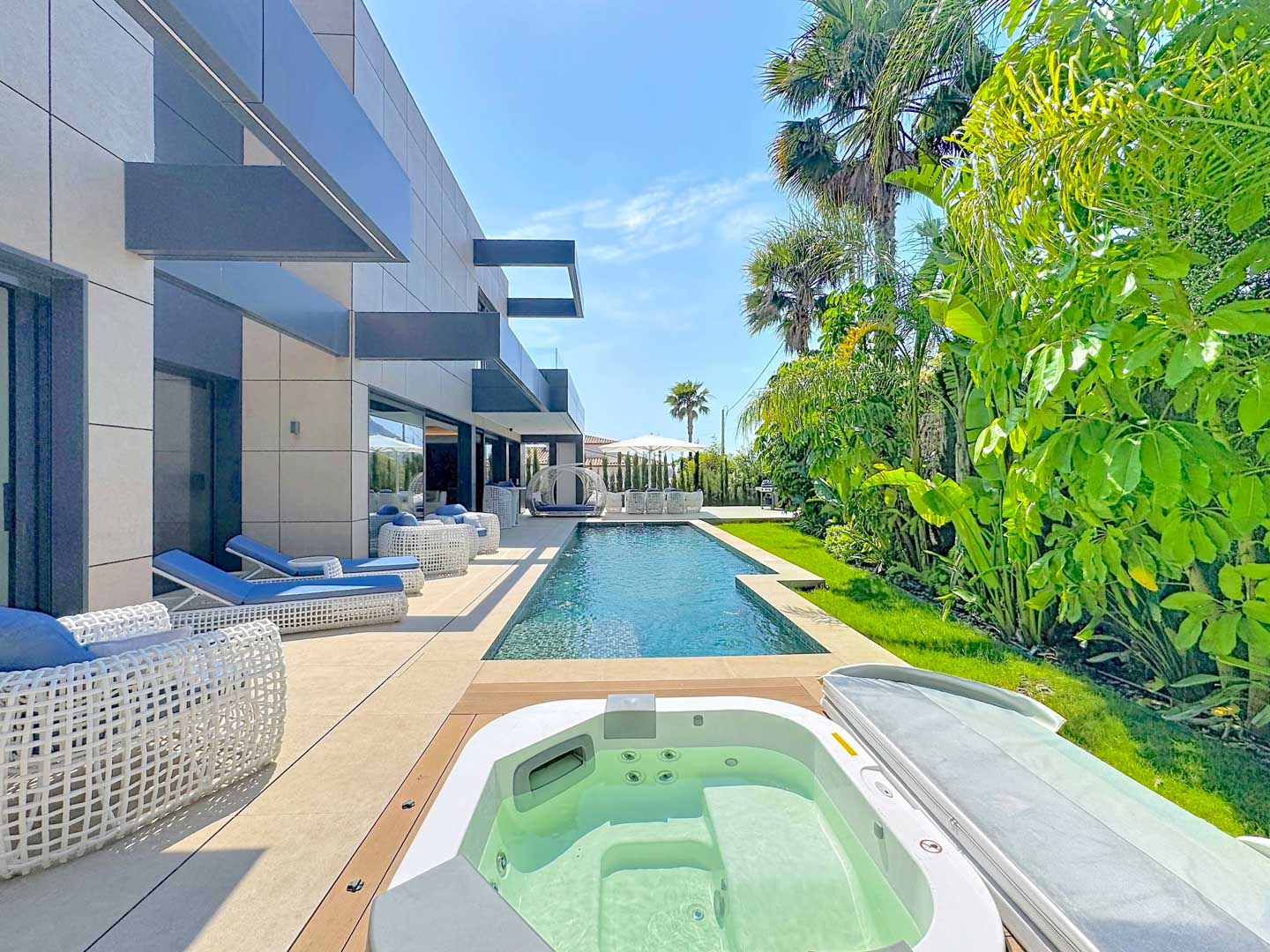 Terraza exterior con piscina rectangular y jacuzzi integrado. Muebles de diseño en blanco y azul complementan la fachada moderna de la villa y el exuberante jardín tropical.