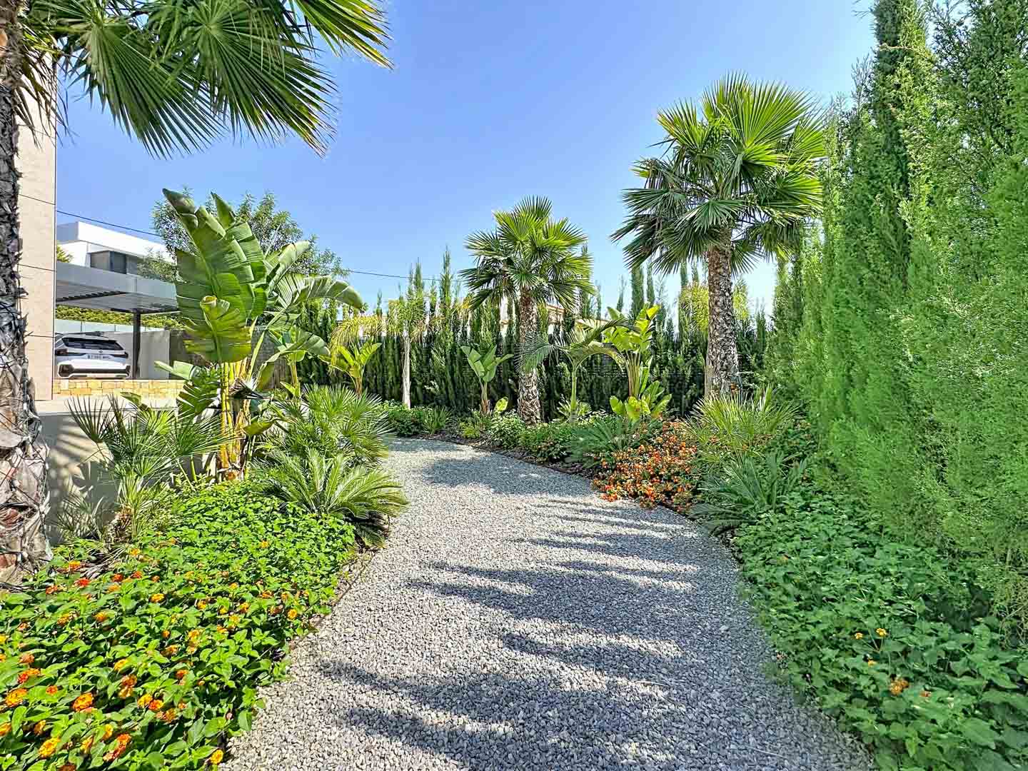 Un sendero serpenteante de grava atraviesa exuberantes jardines tropicales con palmeras variadas, colorida flora y setos de cipreses, hacia la moderna villa y su marquesina.